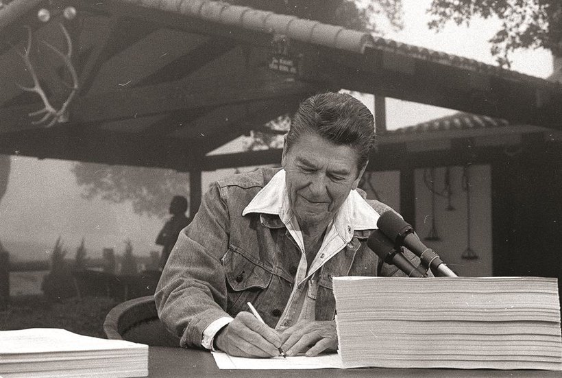 President Ronald Reagan signs the largest tax cut bill in U.S. history at his ranch in California, with the porch and fencing that he built in the background.